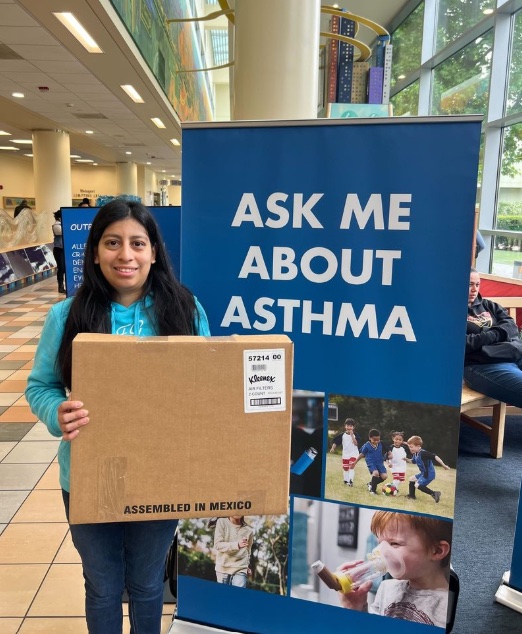 Community member holding a Kleenex® Certified air filters box in front of “Ask Me About Asthma” display.